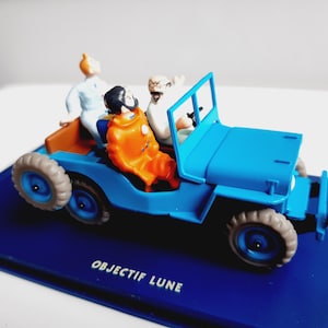 Puede incluir: Un jeep de juguete azul con tres figuras. El jeep tiene carrocer&iacute;a azul, neum&aacute;ticos grises e interior marr&oacute;n. Las figuras son de personajes de dibujos animados, uno con un traje naranja. La base dice "OBJECTIF LUNE".