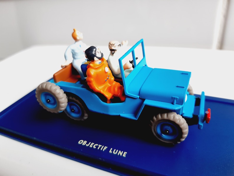 Puede incluir: Un jeep de juguete azul con tres figuras. El jeep tiene carrocer&iacute;a azul, neum&aacute;ticos grises e interior marr&oacute;n. Las figuras son de personajes de dibujos animados, uno con un traje naranja. La base dice "OBJECTIF LUNE".
