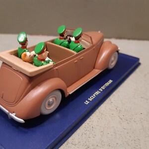 May include: A brown vintage toy car with four figures in green uniforms and hats. The car is on a blue base with the text "LE SCEPTRE D'OTTOKAR". The car has white tires and a tan interior.