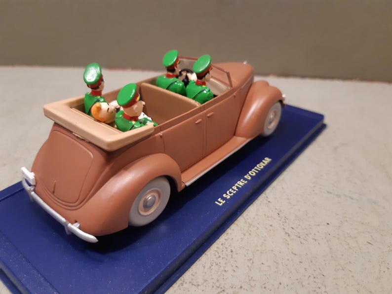 May include: A brown vintage toy car with four figures in green uniforms and hats. The car is on a blue base with the text "LE SCEPTRE D'OTTOKAR". The car has white tires and a tan interior.