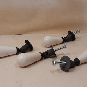 Puede incluir: Cinco pomos de armario de cerámica blanca con detalles negros y tornillos plateados están dispuestos sobre una superficie clara. Los pomos tienen forma de lágrima y están diseñados para muebles. El hardware está listo para la instalación.