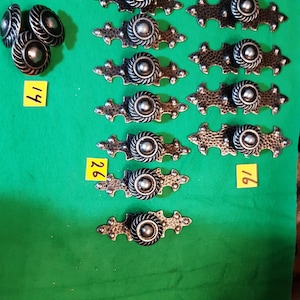 May include: A selection of antique cabinet hardware, including round knobs and decorative pulls with a dark, aged finish. The knobs are stacked, and the pulls are arranged in rows. The background is a solid green. The numbers 14, 26, and 16 are visible.
