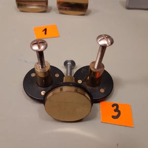 Może przedstawiać: Zbliżenie element&oacute;w sprzętowych, w tym element&oacute;w mosiężnych i czarnego metalu. Na zdjęciu widoczne są trzy okrągłe czarne metalowe podstawy z mosiężnymi śrubami i centralnym mosiężnym dyskiem. Widoczne są r&oacute;wnież pomarańczowe etykiety z numerami 1 i 3.