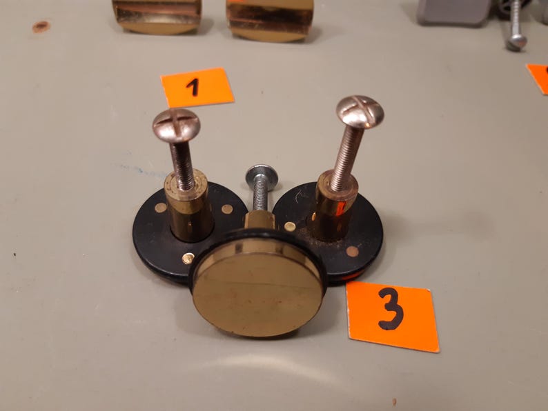 Może przedstawiać: Zbliżenie element&oacute;w sprzętowych, w tym element&oacute;w mosiężnych i czarnego metalu. Na zdjęciu widoczne są trzy okrągłe czarne metalowe podstawy z mosiężnymi śrubami i centralnym mosiężnym dyskiem. Widoczne są r&oacute;wnież pomarańczowe etykiety z numerami 1 i 3.