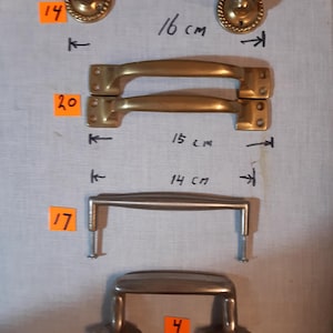 May include: An assortment of vintage metal door and drawer handles. The handles vary in style, including a brass handle measuring 16 cm (6.3 inches) long, and a brushed metal handle measuring 14 cm (5.5 inches) long. The handles are displayed on a light background.