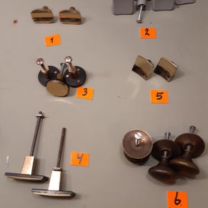 Op de afbeelding: Assortiment van kastbeslag, waaronder gouden en messing knoppen en handgrepen. De collectie bevat verschillende vormen en maten, waaronder vierkante, ronde en rechthoekige ontwerpen. Sommige stukken hebben een geborstelde metalen afwerking, andere zijn gepolijst.