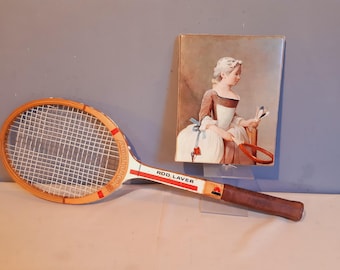 May include: Vintage wooden tennis racket with a white and red handle, and the text "Rod Laver". The racket has a brown leather grip and a white net. Beside it is a framed painting of a woman holding a tennis racket.