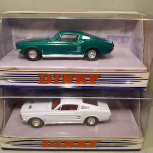 May include: Two Dinky toy cars in clear display cases. One is green, the other white. Both are classic Mustang models. The boxes have the Dinky logo in red and gold. The cars are displayed on a white surface.