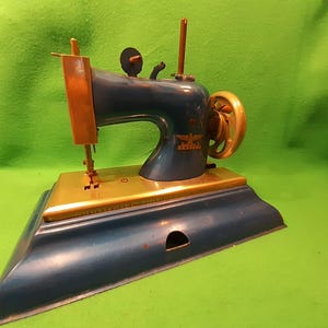 Puede incluir: Máquina de coser vintage azul y dorada. La máquina tiene detalles de latón, incluyendo el volante y el brazo de la aguja. El cuerpo es azul oscuro, y la base también es azul. Cuenta con un emblema decorativo.