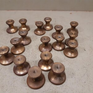 Könnte beinhalten: Eine Sammlung von siebzehn kleinen, runden Metall-Schrankknöpfen mit einem warmen, gebürsteten Bronzefinish. Jeder Knopf verfügt über einen ausgestellten Sockel, einen verjüngten Hals und eine flache Oberseite mit einem mittigen Gewindeloch.