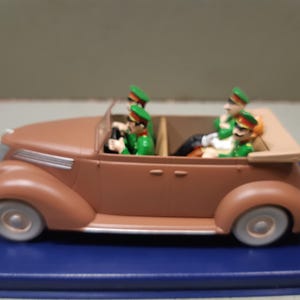 May include: A brown toy convertible car with four figures in green uniforms and hats. The car has white tires and a tan top. The figures appear to be driving and riding in the car.