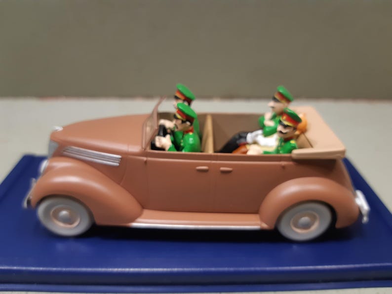 May include: A brown toy convertible car with four figures in green uniforms and hats. The car has white tires and a tan top. The figures appear to be driving and riding in the car.