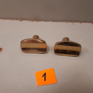 Może przedstawiać: Dwa mosiężne gałki do szafek o prostokątnym kształcie i centralnym poziomym pasku. Gałki mają zaokrągloną g&oacute;rę i cylindryczny trzpień. Pomarańczowa naklejka z numerem "1" znajduje się na pierwszym planie.