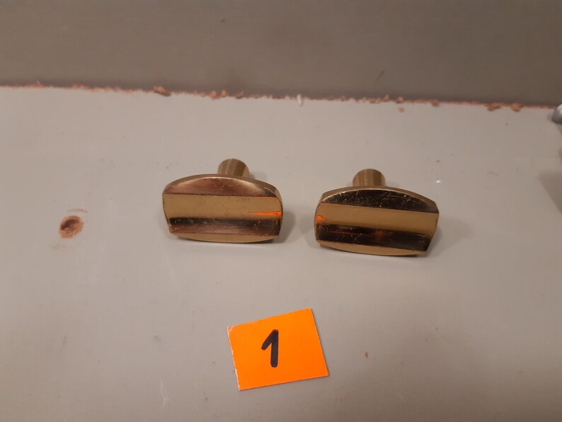 Może przedstawiać: Dwa mosiężne gałki do szafek o prostokątnym kształcie i centralnym poziomym pasku. Gałki mają zaokrągloną g&oacute;rę i cylindryczny trzpień. Pomarańczowa naklejka z numerem "1" znajduje się na pierwszym planie.