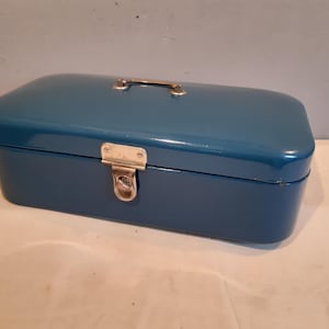 May include: A teal-colored metal toolbox with a rectangular shape. It features a gold-colored handle and a latch closure. The toolbox is likely used for storing tools or other small items.