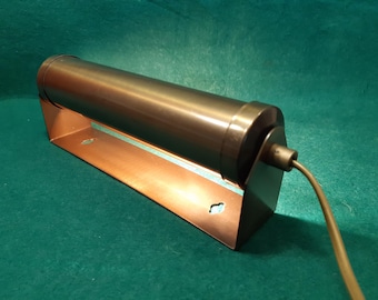 May include: A vintage-style, cylindrical lamp with a dark bronze finish. The lamp is mounted on a copper-colored base with a rectangular shape. A power cord extends from the lamp's side. The background is a solid teal color.