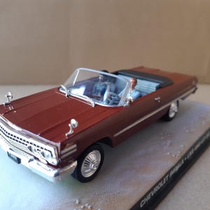 Könnte beinhalten: Ein kastanienbraunes Chevrolet Impala Cabriolet als Modellauto mit verchromtem Kühlergrill, silbernen Radkappen und klarer Windschutzscheibe. Zwei kleine Figuren sitzen im Fahrzeug, das auf einem schwarzen Sockel mit Text steht.