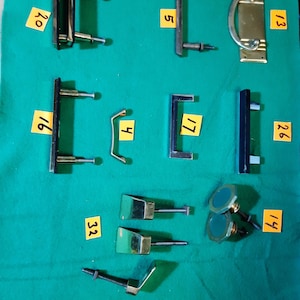 May include: A selection of cabinet hardware, including pulls and knobs, arranged on a green surface. The hardware features various designs and finishes, such as brass, black, and green. Some pieces are rectangular, while others are curved or circular.