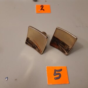 Może przedstawiać: Dwie złote kwadratowe spinki do mankiet&oacute;w o lekko zakrzywionym kształcie. Spinki do mankiet&oacute;w mają błyszczące wykończenie i ciemny obszar u dołu. Tło to jasnoszara powierzchnia z pomarańczowymi etykietami.
