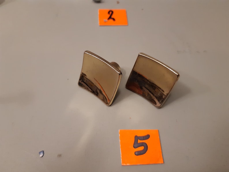 Może przedstawiać: Dwie złote kwadratowe spinki do mankiet&oacute;w o lekko zakrzywionym kształcie. Spinki do mankiet&oacute;w mają błyszczące wykończenie i ciemny obszar u dołu. Tło to jasnoszara powierzchnia z pomarańczowymi etykietami.