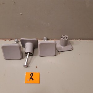 Może przedstawiać: Cztery szare gałki do szafek o kwadratowych powierzchniach i cylindrycznej podstawie. Jedna gałka ma przymocowaną śrubę. Mała pomarańczowa naklejka z numerem "2" jest na pierwszym planie.