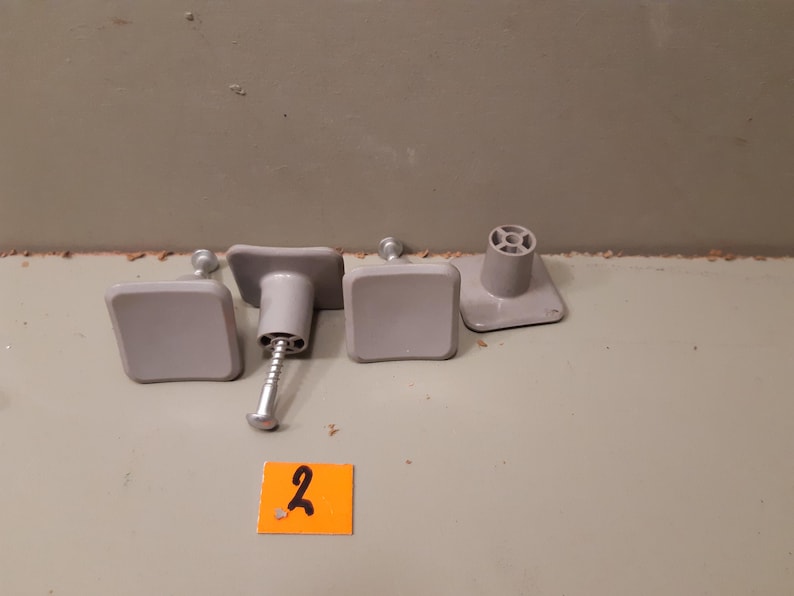 Może przedstawiać: Cztery szare gałki do szafek o kwadratowych powierzchniach i cylindrycznej podstawie. Jedna gałka ma przymocowaną śrubę. Mała pomarańczowa naklejka z numerem "2" jest na pierwszym planie.