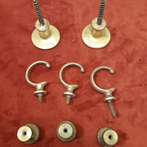 Op de afbeelding: Een assortiment vintage messing hardware, waaronder twee ronde knoppen met schroeven, drie C-vormige haken met schroeven en drie kleinere ronde knoppen. De hardware is geplaatst tegen een rode achtergrond.