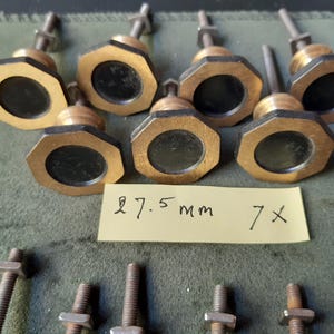 Peut inclure: Sept boutons octogonaux avec une finition dor&eacute;e m&eacute;tallique et un insert circulaire noir au centre. Chaque bouton comporte un boulon m&eacute;tallique filet&eacute; et un &eacute;crou. Une note en papier jaune porte le texte manuscrit 27.5 mm 7x.