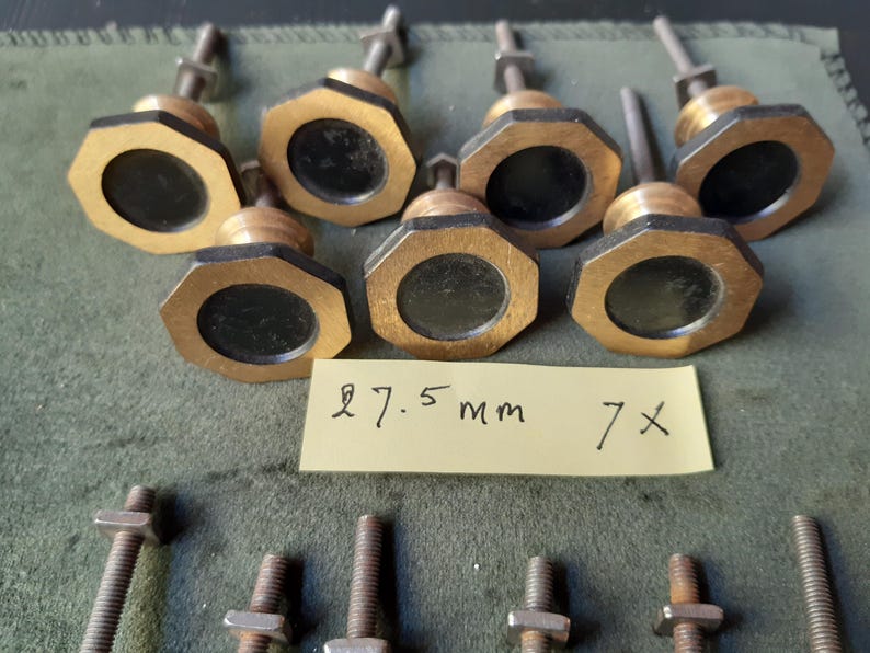Peut inclure: Sept boutons octogonaux avec une finition dor&eacute;e m&eacute;tallique et un insert circulaire noir au centre. Chaque bouton comporte un boulon m&eacute;tallique filet&eacute; et un &eacute;crou. Une note en papier jaune porte le texte manuscrit 27.5 mm 7x.