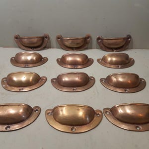 Peut inclure: Ensemble de onze poignées de tiroir antiques en laiton. Les poignées ont une forme en demi-lune avec une cuvette arrondie et une base plate avec des trous de vis. Le laiton présente une patine chaude et vieillie, suggérant du matériel vintage.