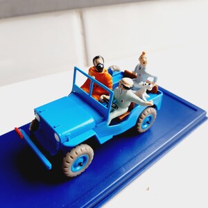 Puede incluir: Un jeep azul en miniatura con tres figuras y un perro. El jeep tiene neum&aacute;ticos color canela y un parachoques rojo. Las figuras son de personas con varios atuendos, incluido un hombre con una t&uacute;nica naranja y un hombre con un traje azul claro.