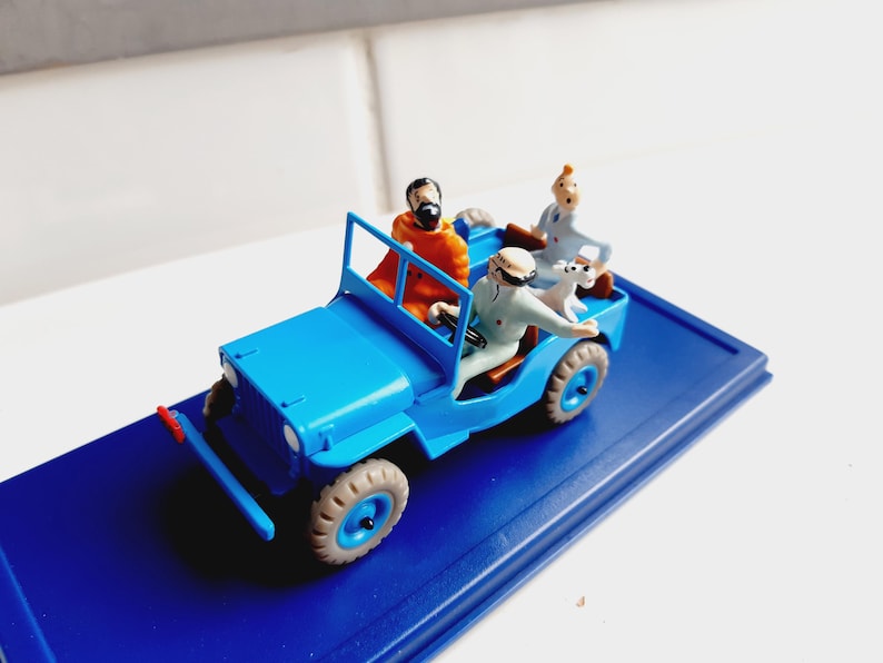 Puede incluir: Un jeep azul en miniatura con tres figuras y un perro. El jeep tiene neum&aacute;ticos color canela y un parachoques rojo. Las figuras son de personas con varios atuendos, incluido un hombre con una t&uacute;nica naranja y un hombre con un traje azul claro.