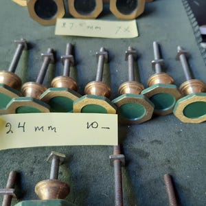 Peut inclure: Une collection de boutons en m&eacute;tal avec des cadres octogonaux couleur laiton et des centres circulaires vert fonc&eacute;. Chaque bouton comporte un boulon filet&eacute; et un &eacute;crou carr&eacute;. Une note adh&eacute;sive jaune porte le texte manuscrit 24 mm 10-.