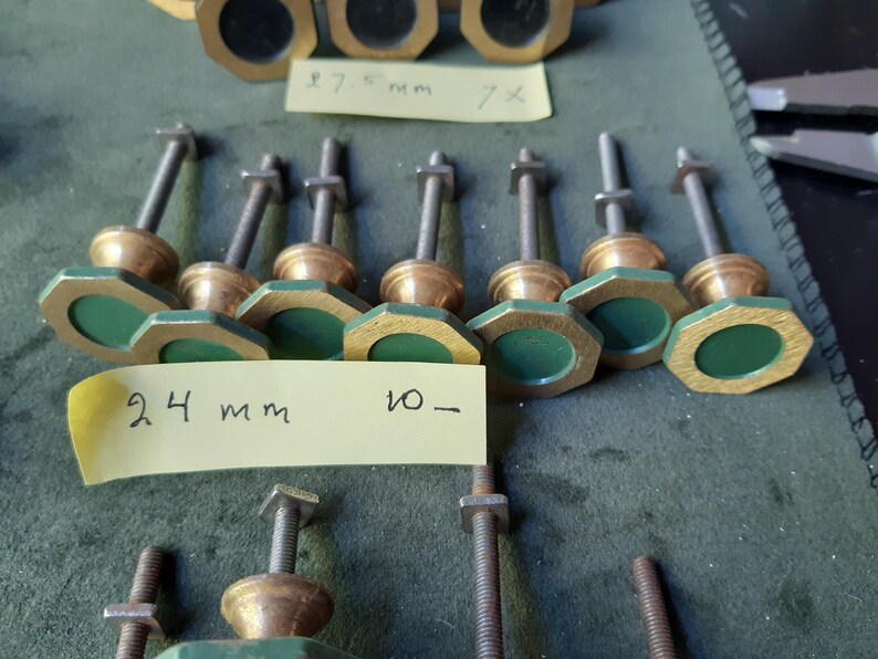 Peut inclure: Une collection de boutons en m&eacute;tal avec des cadres octogonaux couleur laiton et des centres circulaires vert fonc&eacute;. Chaque bouton comporte un boulon filet&eacute; et un &eacute;crou carr&eacute;. Une note adh&eacute;sive jaune porte le texte manuscrit 24 mm 10-.