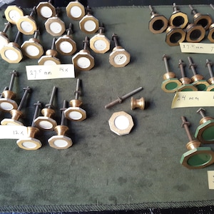 May include: A collection of metal cabinet knobs with octagonal faces in white, black, and green, organized by size. Labels indicate 27.5 mm, 24 mm, and 35 mm measurements. Each knob features a threaded metal post and a decorative metal base.