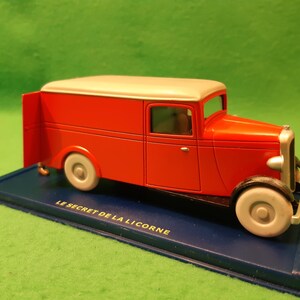 以下が含まれることがあります： クリーム色の屋根と白い車輪が付いた赤いおもちゃの配達トラック。トラックは「LE SECRET DE LA LICORNE」の文字が入った青い台座の上にあります。クラシックなデザインの車です。