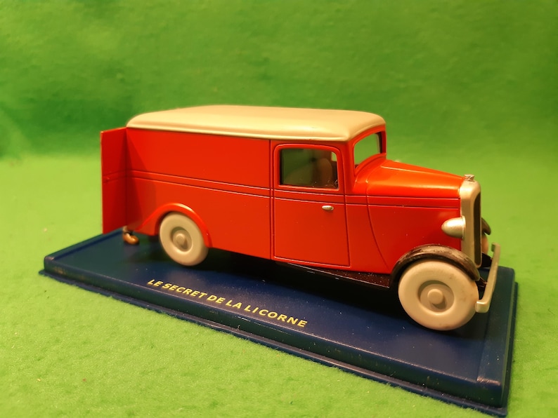 以下が含まれることがあります： クリーム色の屋根と白い車輪が付いた赤いおもちゃの配達トラック。トラックは「LE SECRET DE LA LICORNE」の文字が入った青い台座の上にあります。クラシックなデザインの車です。