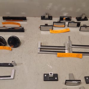 Op de afbeelding: Een verzameling van diverse metalen en zwarte kastgrepen en knoppen, inclusief rechthoekige en ronde ontwerpen. Verschillende onderdelen zijn voorzien van oranje plakbriefjes met daarop maten zoals 90 mm, 42 mm, 36 mm en 60 mm.