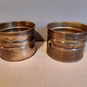Op de afbeelding: Twee antieke messing plantenbakken met een gehamerde textuur en ringgrepen. De plantenbakken hebben een cilindrische vorm met een licht uitlopende rand. Het messing heeft een warme, verouderde patina.
