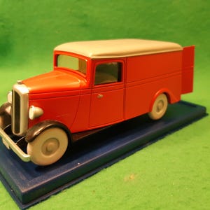以下が含まれることがあります： クリーム色の屋根と白い車輪が付いた赤いヴィンテージの配送トラックモデル。トラックは青いベースにあり、フェンダーとグリルに黒いアクセントがあります。配送車のクラシックな表現です。