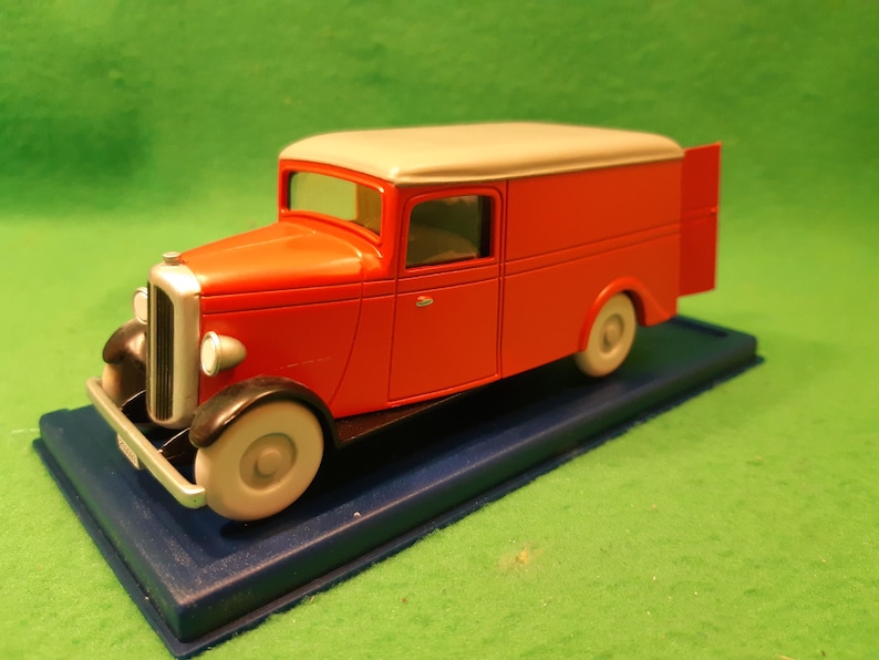 以下が含まれることがあります： クリーム色の屋根と白い車輪が付いた赤いヴィンテージの配送トラックモデル。トラックは青いベースにあり、フェンダーとグリルに黒いアクセントがあります。配送車のクラシックな表現です。