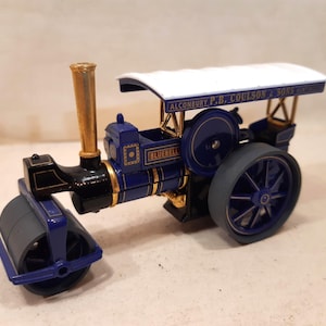 Op de afbeelding: Een miniatuur blauwe en zwarte stoomwals met gouden accenten en een wit dak. De speelgoedwagen heeft de tekst "BLUEBELL" en "ALCONBURY P.B. COULSON & SONS" op de carrosserie. Een klassiek speelgoed of verzamelobject.