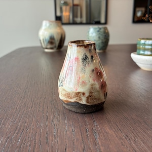 Könnte beinhalten: Kleine, handgefertigte Keramikvase mit einer einzigartigen, organischen Form. Die Vase hat einen braunen Sockel, einen cremefarbenen Körper mit rosa und schwarzen Akzenten und einen braunen Rand. Weitere Keramikteile sind im Hintergrund.