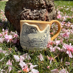 May include: A ceramic mug with a tan and cream colour scheme. The mug features a black and white illustration of an animal holding a tea tray. The mug is sitting in grass with pink flower petals.