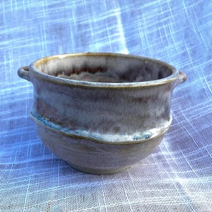 May include: A handmade ceramic bowl with a wide opening and two small handles. The bowl features a blend of colors, including brown, gray, and a touch of blue. The rim has a golden hue.