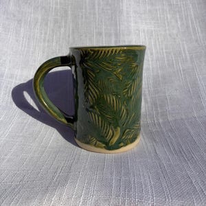 Könnte beinhalten: Grüne Keramiktasse mit Blattmuster. Die Tasse hat einen abgerundeten Henkel und einen hellen Boden. Die Außenseite ist tiefgrün mit einem strukturierten Blattdesign, ideal für Tee oder Kaffee.