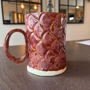 May include: A handmade ceramic mug with a textured, scale-like pattern in shades of red and brown. The mug has a large, rounded handle and a white base. The mug is sitting on a wooden table.