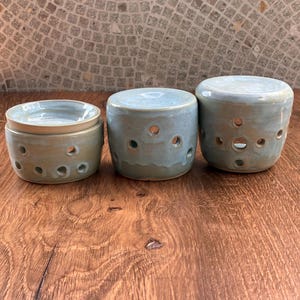 May include: Three cylindrical ceramic wax warmers with a glossy light blue glaze. Each piece features a series of small circular cutouts around the base. The leftmost warmer includes a separate shallow dish on top, while the others are solid.