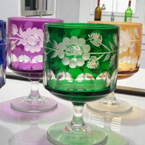 May include: A collection of colorful, etched glass goblets. The goblets are in shades of purple, green, and yellow, each featuring a floral design. The stems and bases are clear, and the glasses are arranged on a reflective surface.