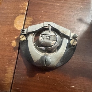 May include: A vintage sewing machine bobbin case, featuring a silver-toned metal frame with a circular design. The bobbin case is set against a dark wooden surface, showcasing its intricate mechanical details and aged appearance.